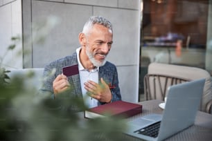Anciano enérgico sosteniendo una tarjeta de plástico rojo y gesticulando con la mano frente a la computadora portátil