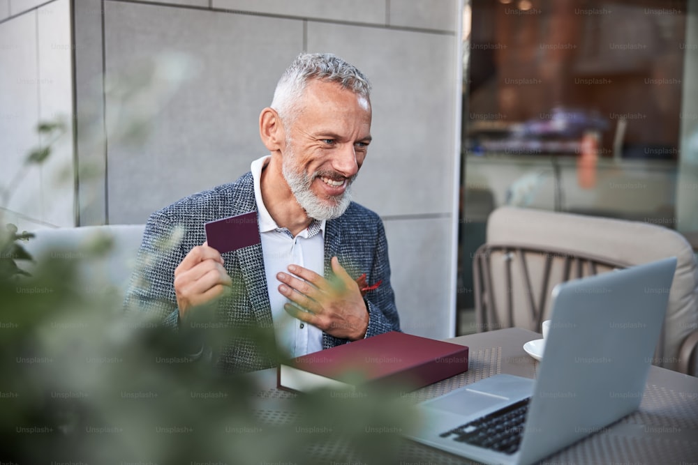 Energetic aging man holding a dar-red plastic card and gesturing with his hand in front of laptop