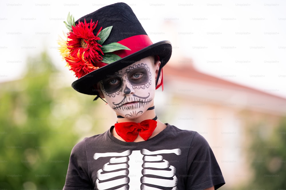 Menino assustador com rosto pintado em halloween fantasia de esqueleto com chapéu preto elegante em pé na frente da câmera e olhando para você