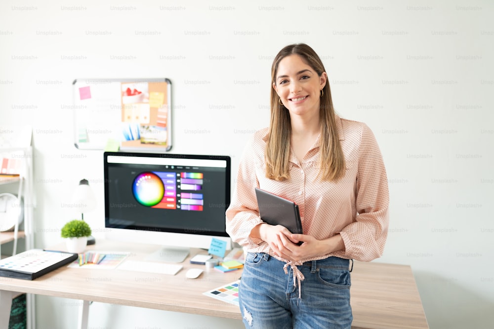 Good looking Caucasian illustrator holding a graphic tablet while standing next to her desk in her office