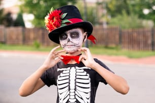 Halloween-Junge mit bemaltem Gesicht im Kostüm des Skeletts mit elegantem Hut und roter Fliege am Hals, der vor der Kamera gegen die Straße steht