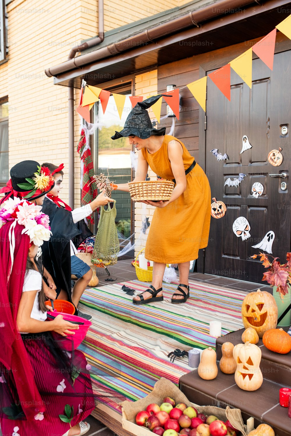 Young cheerful woman in yellow dress and black witch hat bending over staircase by group of halloween kids while giving them sweet treats