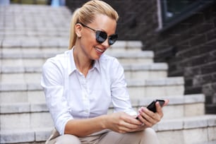Donna bionda alla moda attraente e affascinante con occhiali da sole seduta sulle scale all'aperto e usando lo smartphone.