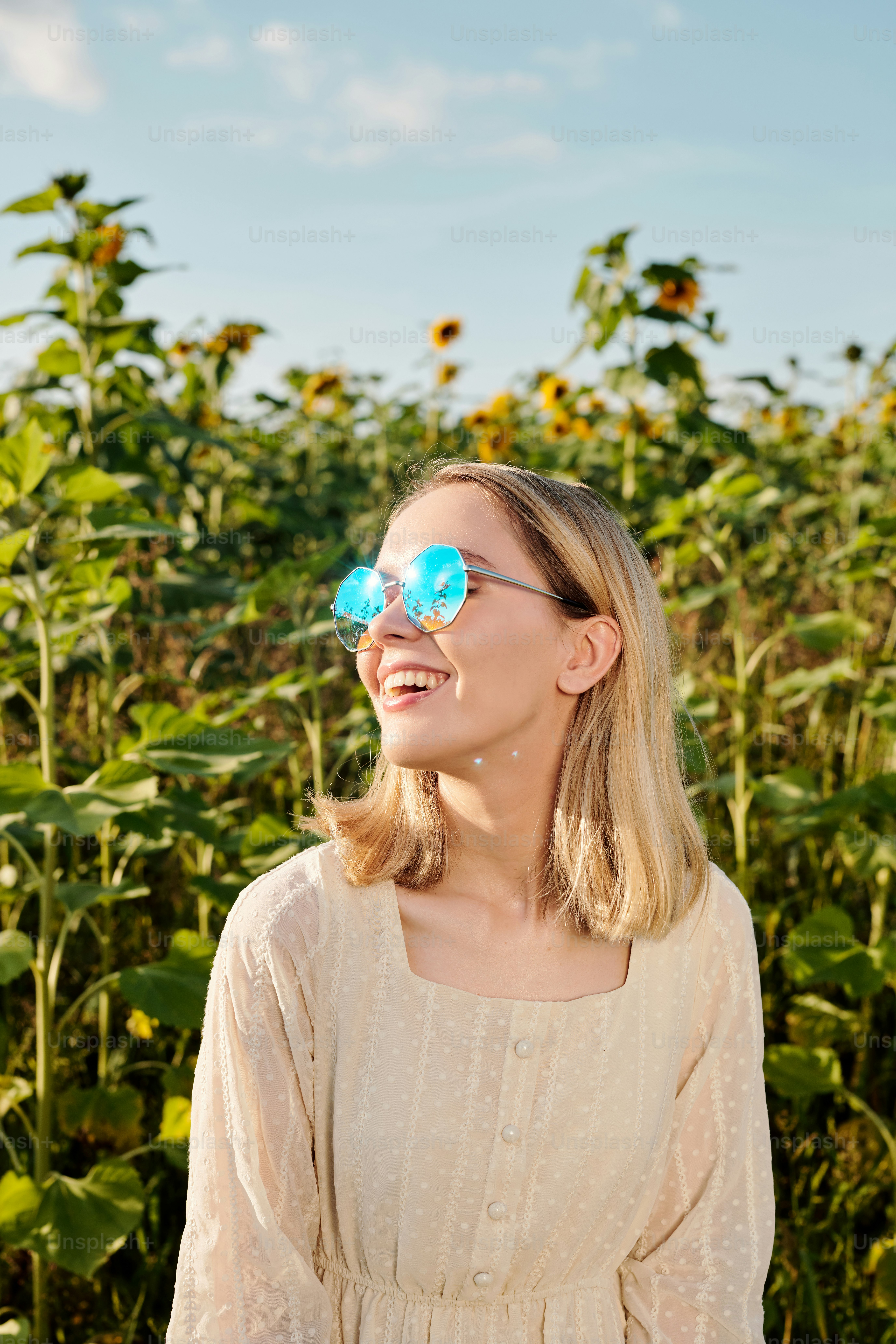 Mulher loira jovem alegre em óculos escuros e vestido branco de pé contra o campo de girassol na frente da câmera com céu azul no fundo