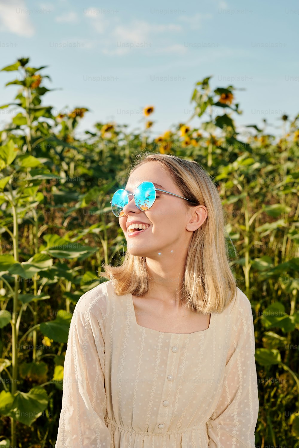 Joyful young blond woman in sunglasses and white dress standing against sunflower field in front of camera with blue sky on background