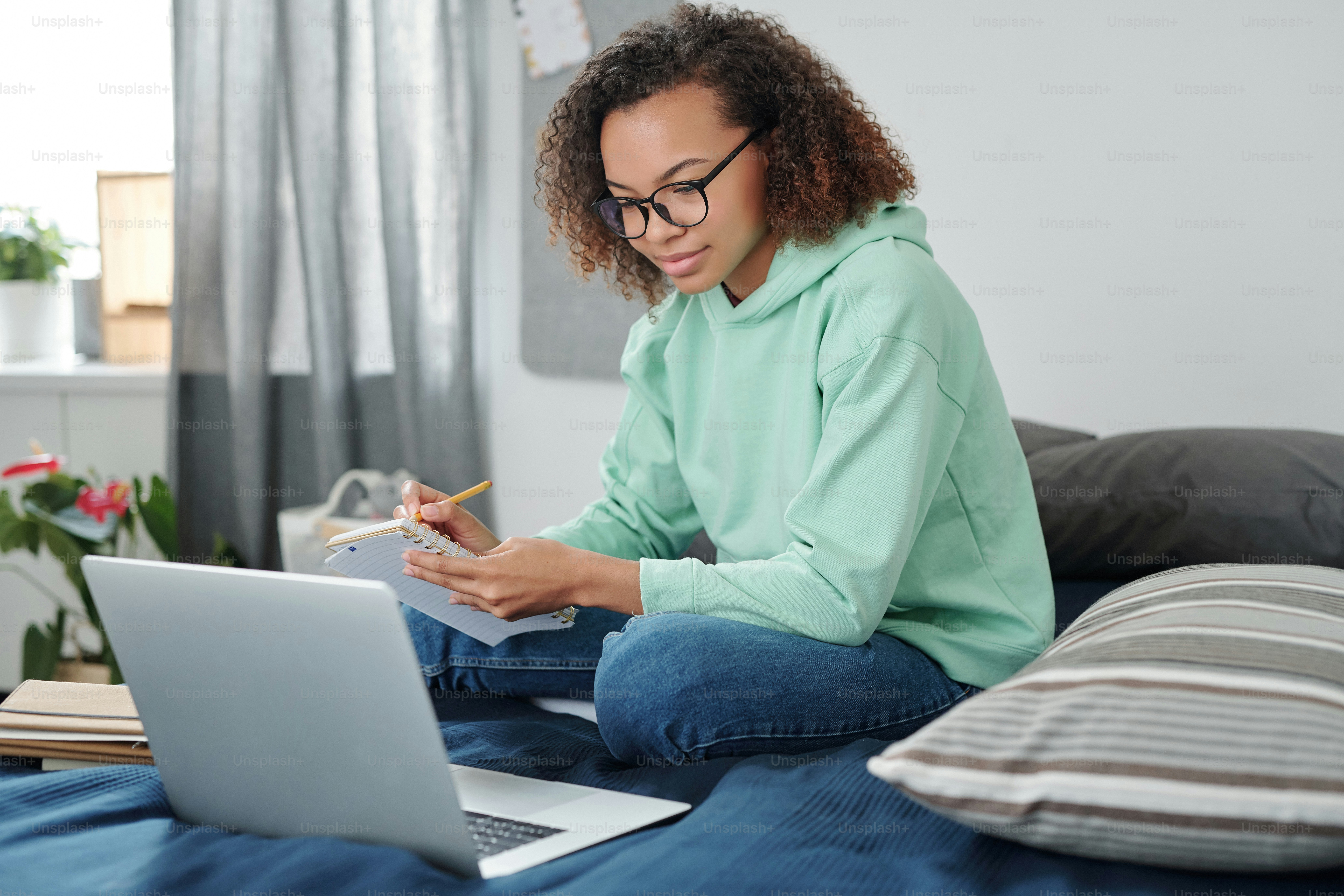 Siswa perempuan ras campuran muda dengan pakaian kasual duduk di tempat tidur di depan laptop, membuat catatan dan mendengarkan guru selama pelajaran online