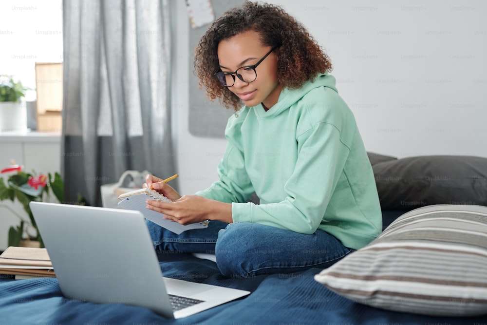 Young mixed-race female student in casualwear sitting on bed in front of laptop, making notes and listening to teacher during online lesson