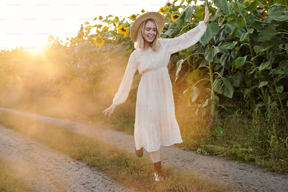 帽子とドレスを着た陽気な金髪の女の子が、晴れた朝に田舎の環境で楽しみながら、ひまわりの間の田舎道を移動する