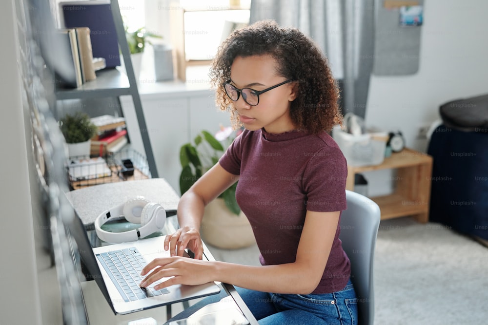 Estudante jovem contemporânea em casualwear sentada por pequena mesa na frente do laptop enquanto faz curso on-line de estudo no quarto