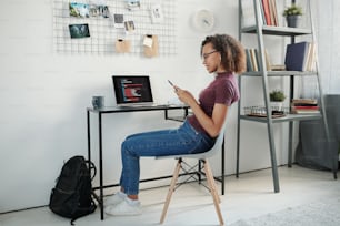 Jeune étudiante métisse joyeuse en tenue décontractée et écouteurs assise près d’une petite table devant un ordinateur portable tout en faisant défiler son smartphone