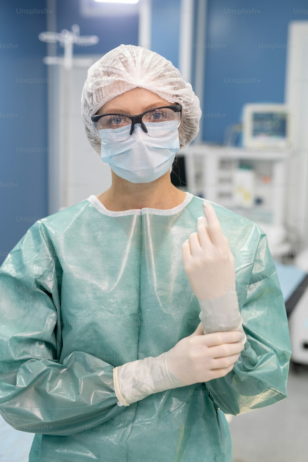 보호복, 장갑, 마스크, 안경, 작업복을 입은 젊은 전문 여성 외과 의사가 수술실에서 카메라 앞에 서 있다