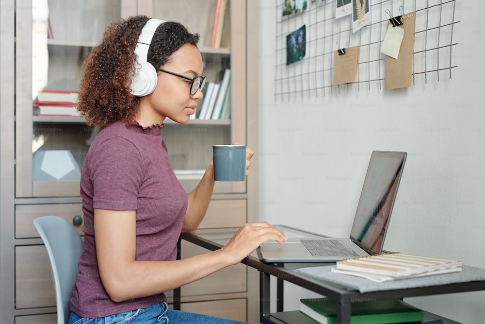 Jovem estudante mestiça em roupas casuais e fones de ouvido sentada na frente do laptop enquanto toma chá e faz networking no ambiente doméstico
