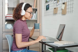 Junge gemischtrassige Studentin in Freizeitkleidung und Kopfhörern, die vor dem Laptop sitzt, während sie Tee trinkt und sich in der häuslichen Umgebung vernetzt