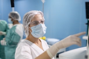 Junge Assistentin des Chirurgen in Handschuhen, Maske und Uniform zeigt auf die Auslage medizinischer Geräte und übernimmt dabei die Kontrolle über die Patientendaten