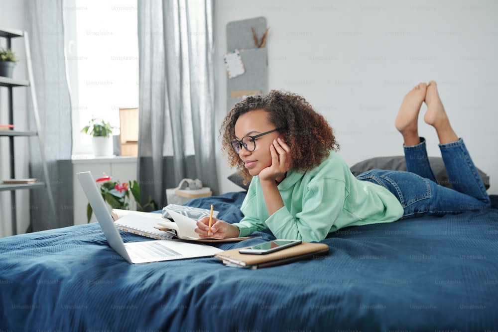 Young clever mixed-race student in jeans and hoodie lying on bed in front of laptop and watching online lesson while making notes in copybook