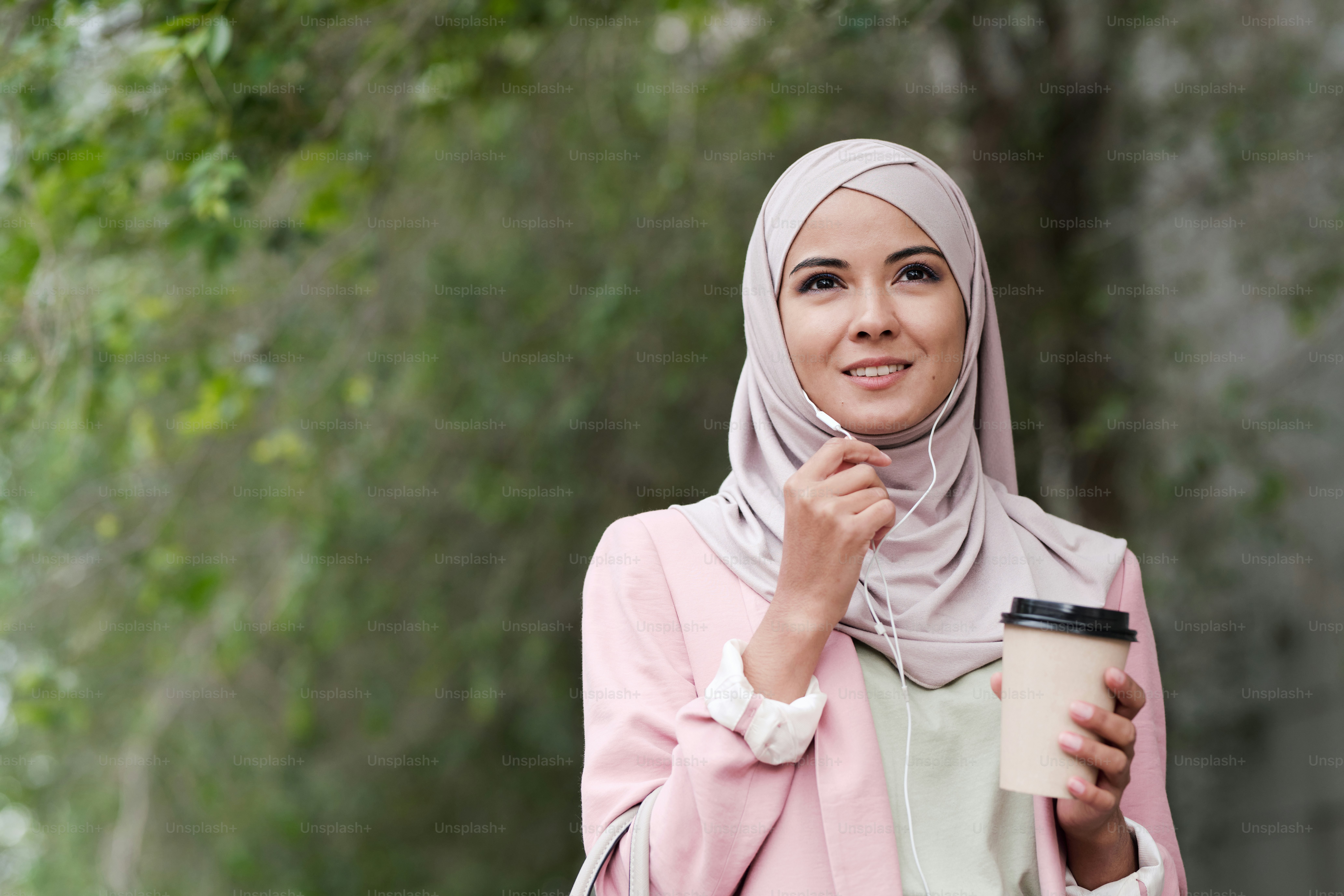 Jovem mulher muçulmana linda em fones de ouvido tomando café e falando com alguém ao telefone enquanto está em pé no ambiente urbano