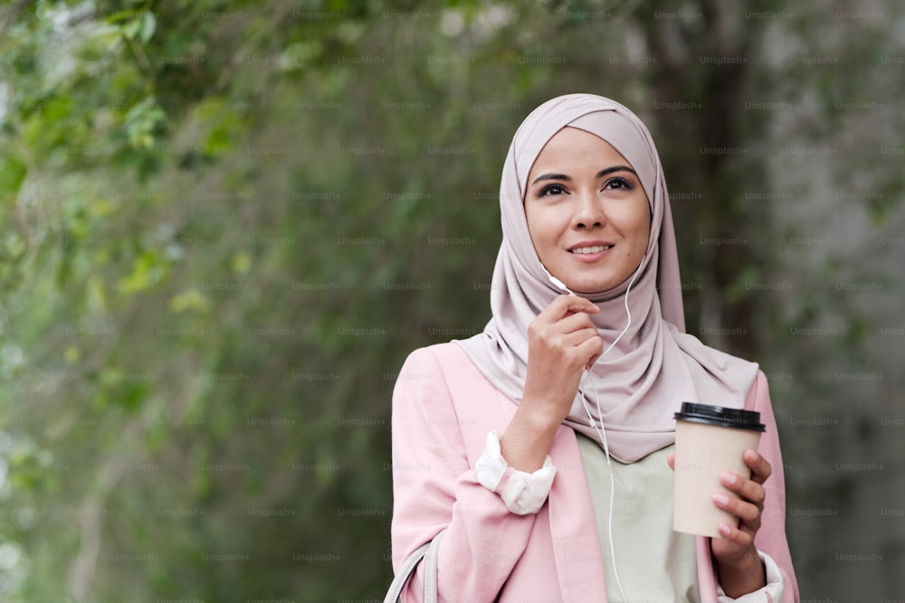 Joven y hermosa mujer musulmana con auriculares tomando café y hablando con alguien por teléfono mientras está de pie en un entorno urbano