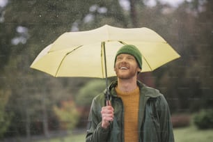 Under umbrella. Young bearded man standing with yellow umbrella in hand