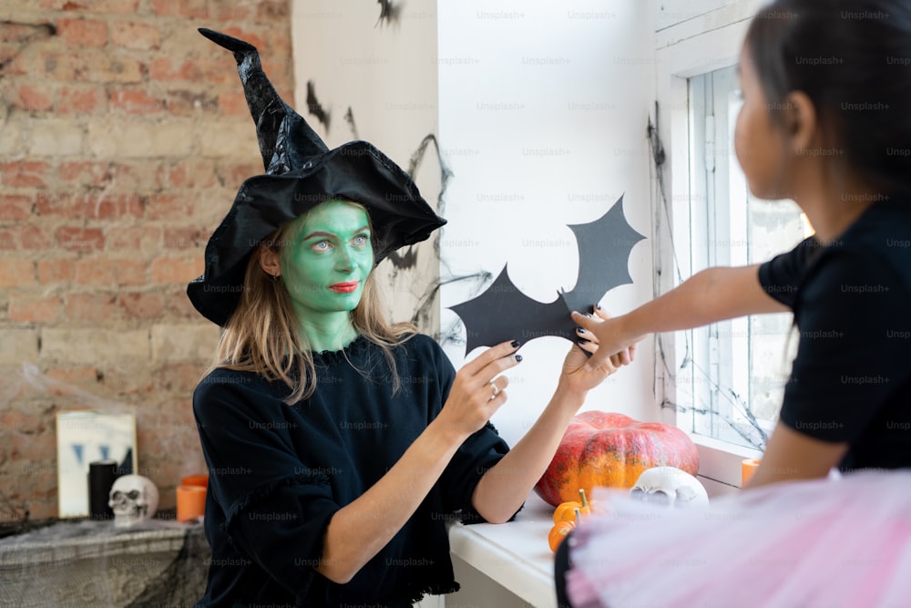Witch woman with green makeup giving paper rat to girl while they decorating room together for Halloween