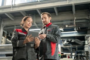 Dos jóvenes gerentes de control de calidad de una fábrica contemporánea de procesamiento de polímeros mirando la pantalla del panel táctil mientras miran los datos