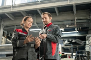 Two young quality control managers of contemporary polymer processing factory looking at touchpad display while looking through data