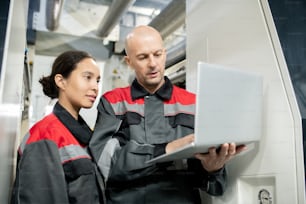 Selbstbewusster, glatzköpfiger, reifer Vorarbeiter in Arbeitskleidung, der einen Laptop hält, während er jungen weiblichen Untergebenen neue Materialien vorstellt