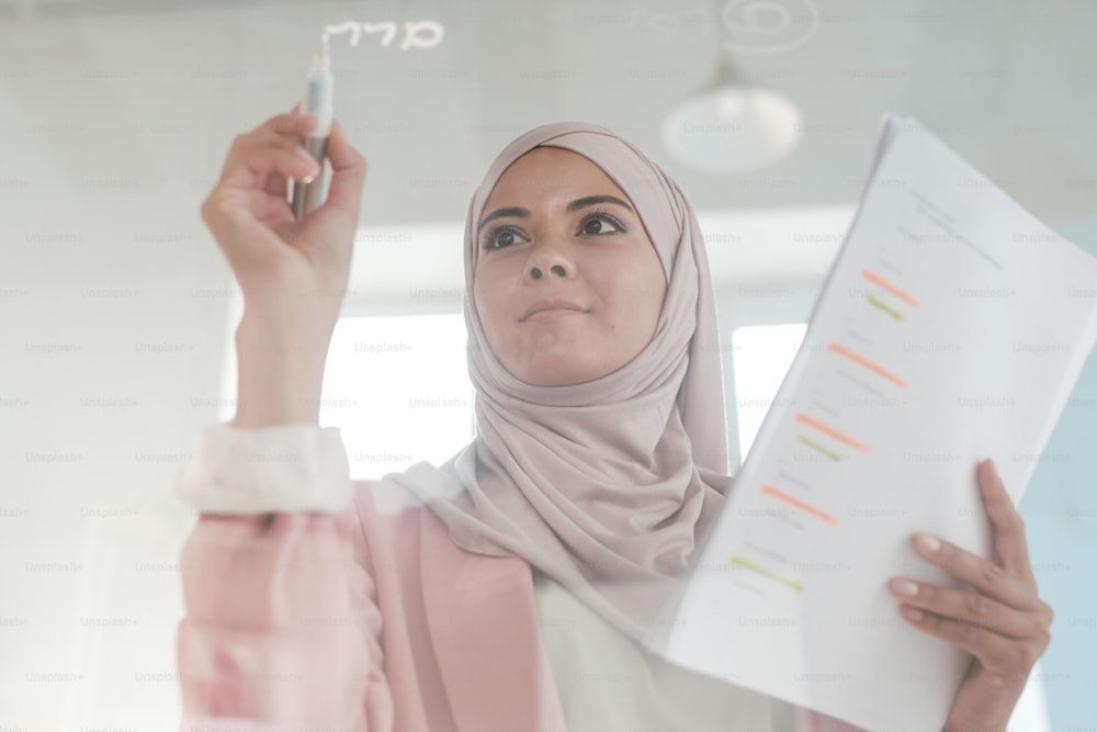 Joven corredora seria con hijab sosteniendo papeles financieros y resaltador mientras hace una presentación por parte de un tablero transparente