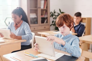Ernsthafter Schüler mit Touchpad, der Online-Dokumente durchblättert, während er während der Einzelarbeit am Schreibtisch gegen interkulturelle Klassenkameraden sitzt