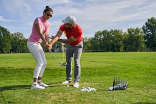 Ernsthafte Golfanfängerin mit Sonnenbrille, die lernt, einen Schläger zu halten, unterstützt von ihrem persönlichen Trainer