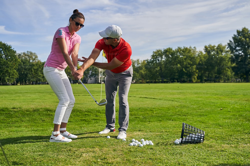 Serious beginner golfer in sunglasses learning to hold a club assisted by her personal coach