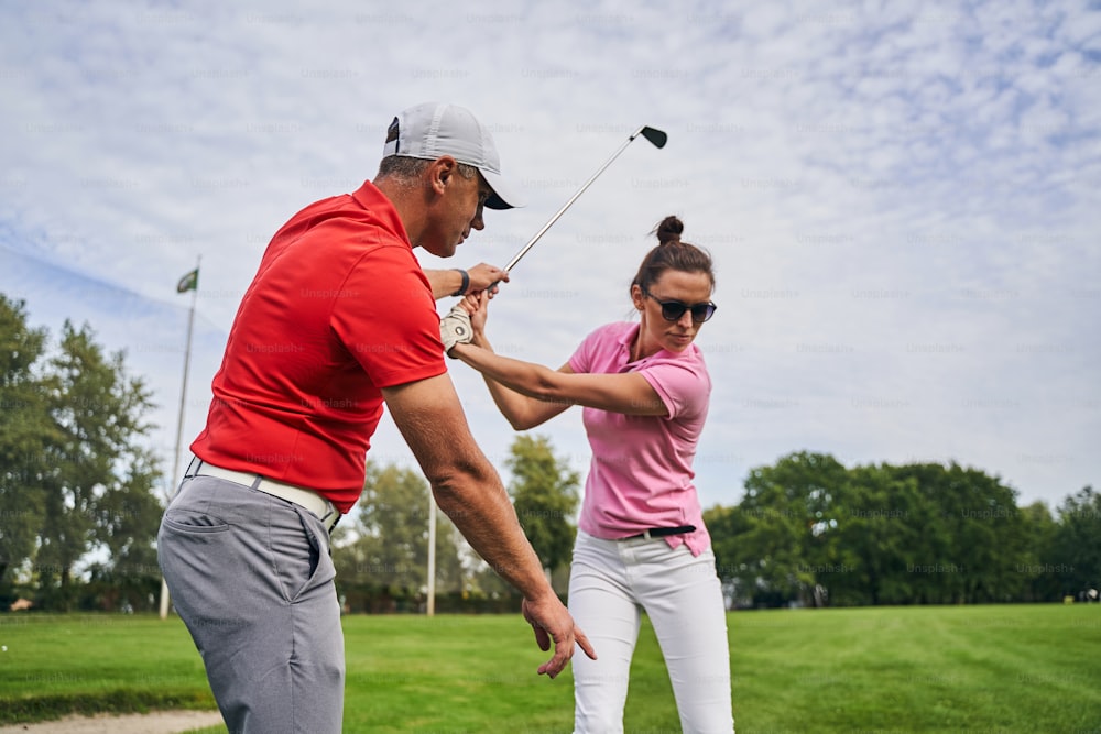 Skilled middle-aged male instructor in a cap explaining the golf fundamentals to a female beginner