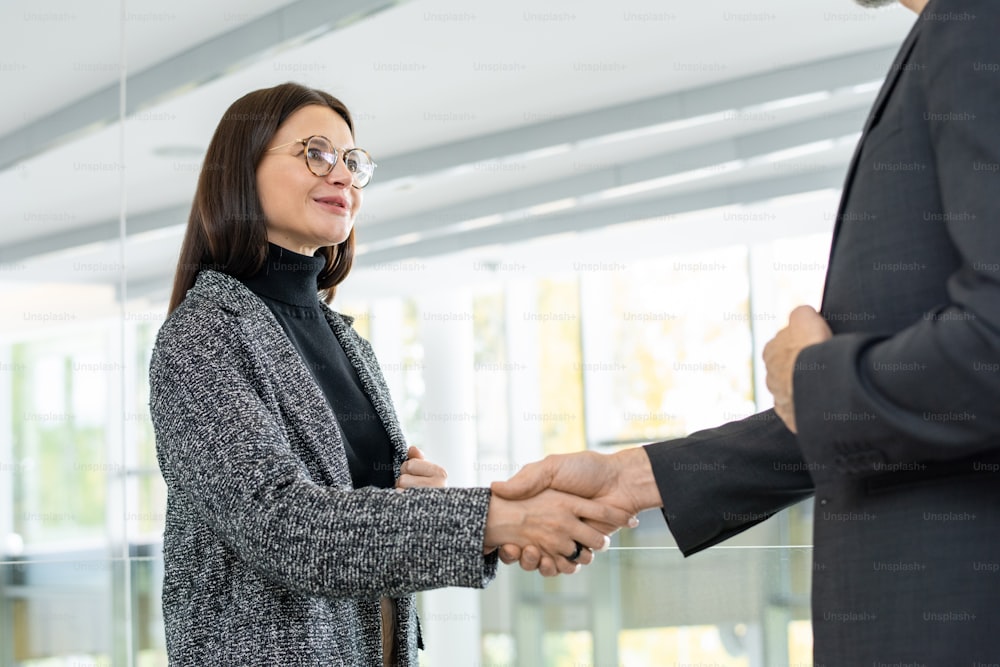 Contemporary young successful businesswoman shaking hand of her male business partner or director of company in office against window