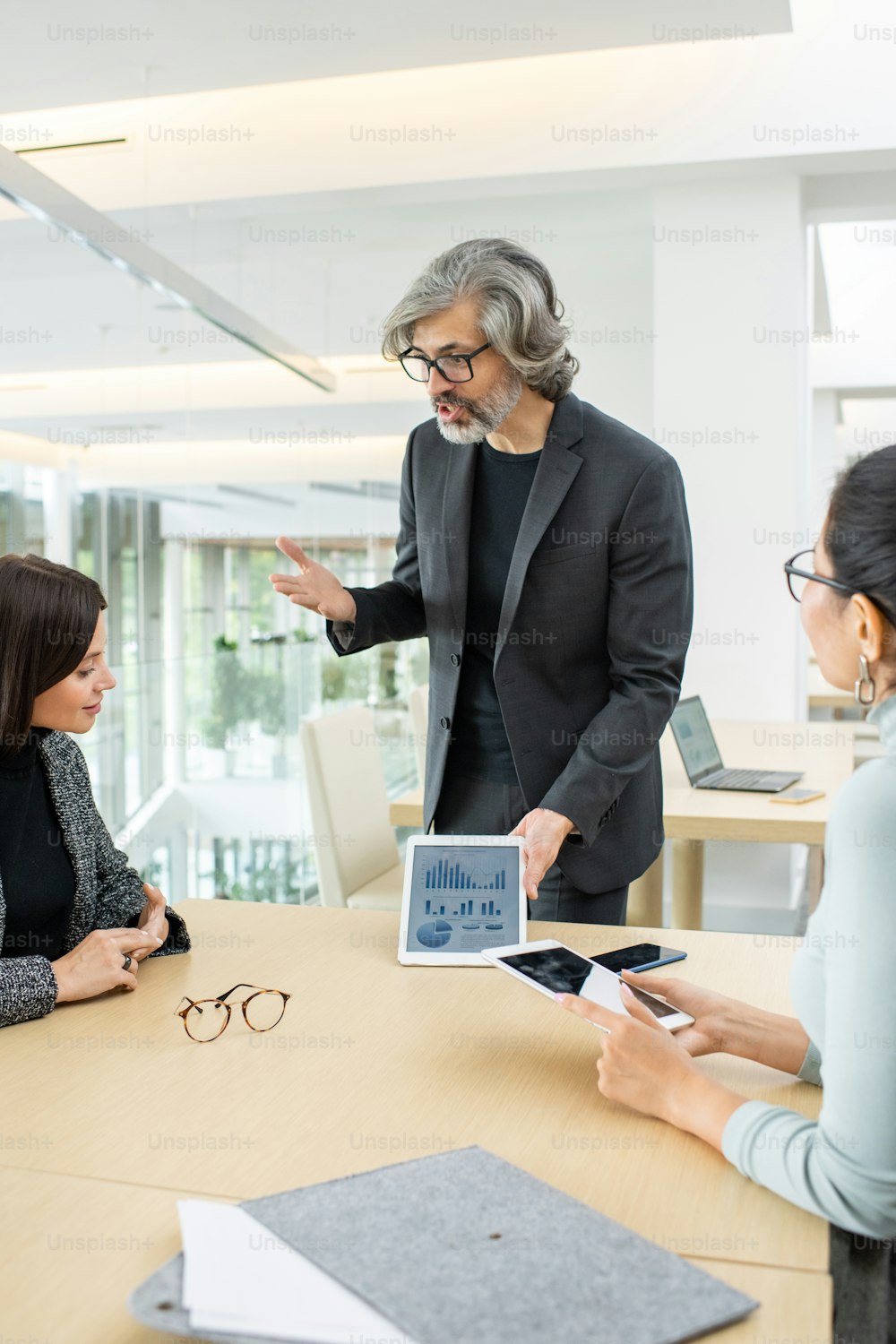 Économiste masculin mature et confiant avec pavé tactile debout près de la table et expliquant les données financières à ses jeunes collègues féminines lors d’une réunion