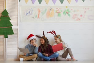 Full length portrait of multi-ethnic group of kids sitting on floor holding pictures while enjoying art class on Christmas, copy space