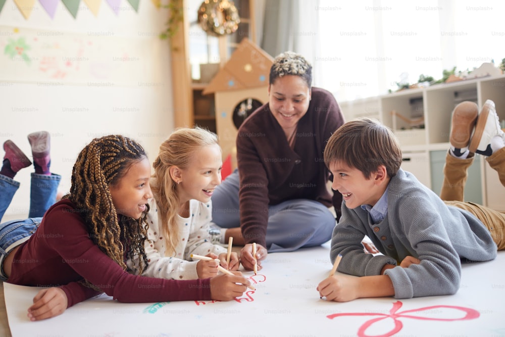 Full length portrait of smiling female teacher sitting on floor with multi-ethnic group of kids drawing pictures while enjoying art class, copy space