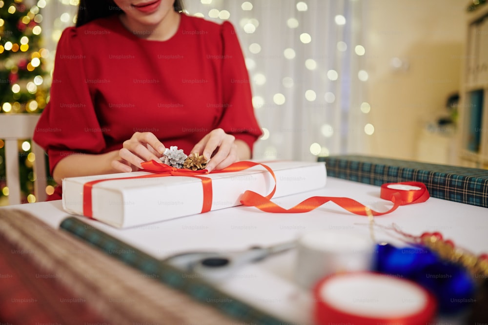 Cropped image of young woman in red dress decorating Christmas present with pine cone