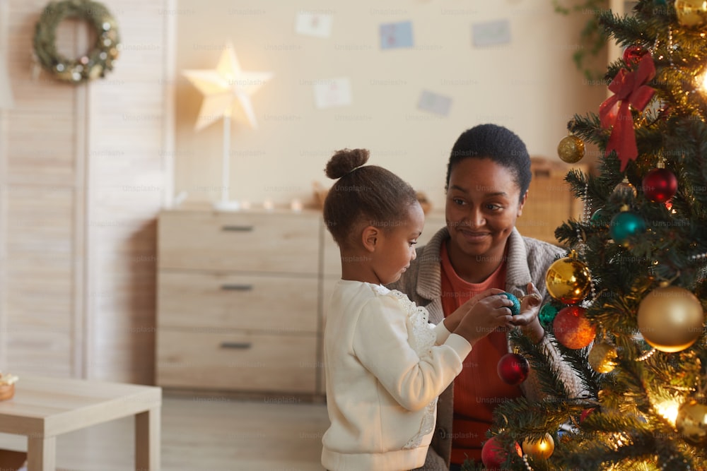 Retrato da vista lateral da menina afro-americana bonito decorando a árvore de Natal com a mãe feliz sorridente no interior aconchegante da casa, espaço da cópia