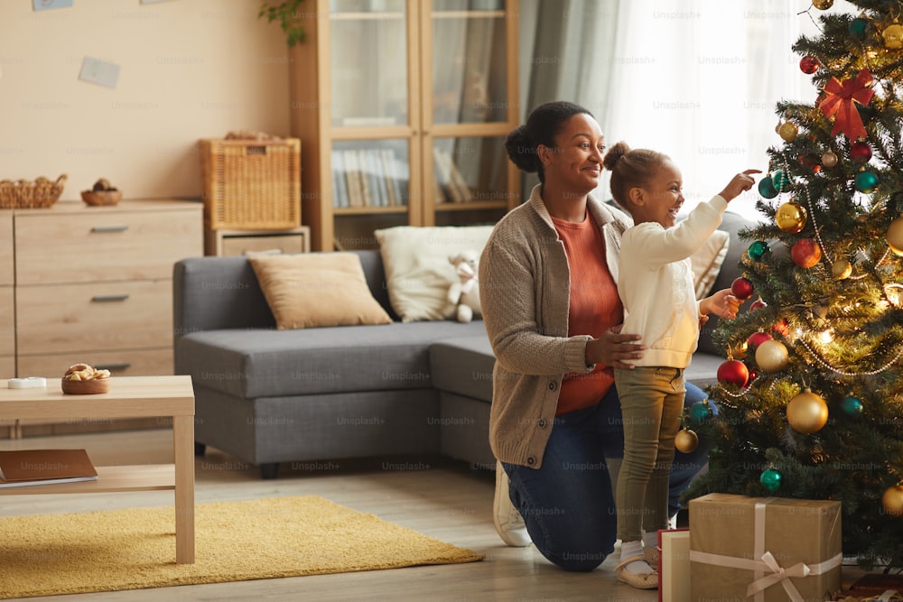 Retrato de vista lateral de comprimento total da menina afro-americana bonito decorando a �árvore de Natal com a mãe feliz sorridente no interior aconchegante da casa, espaço de cópia