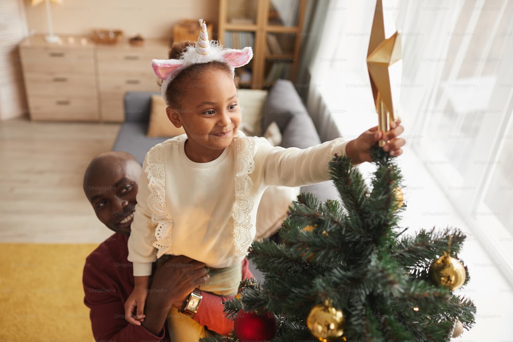 Hochwinkelporträt eines niedlichen afroamerikanischen Mädchens, das einen Stern auf den Weihnachtsbaum setzt, mit einem liebevollen Vater, der ihr hilft, während sie die Weihnachtszeit zu Hause genießt
