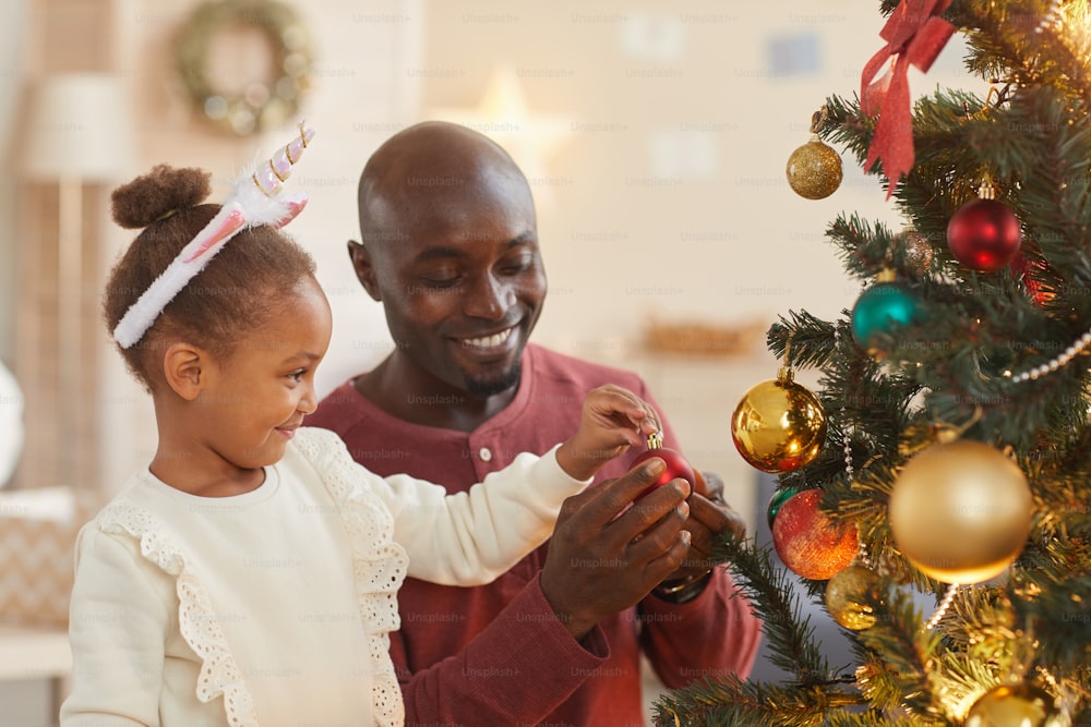 Retrato da menina afro-americana bonito decorando a árvore de Natal com o pai amoroso enquanto desfruta da temporada de férias em casa