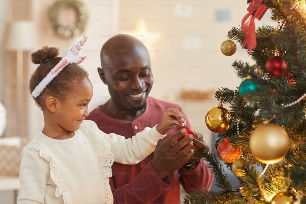 Portrait of cute African-American girl decorating Christmas tree with loving father while enjoying holiday season at home