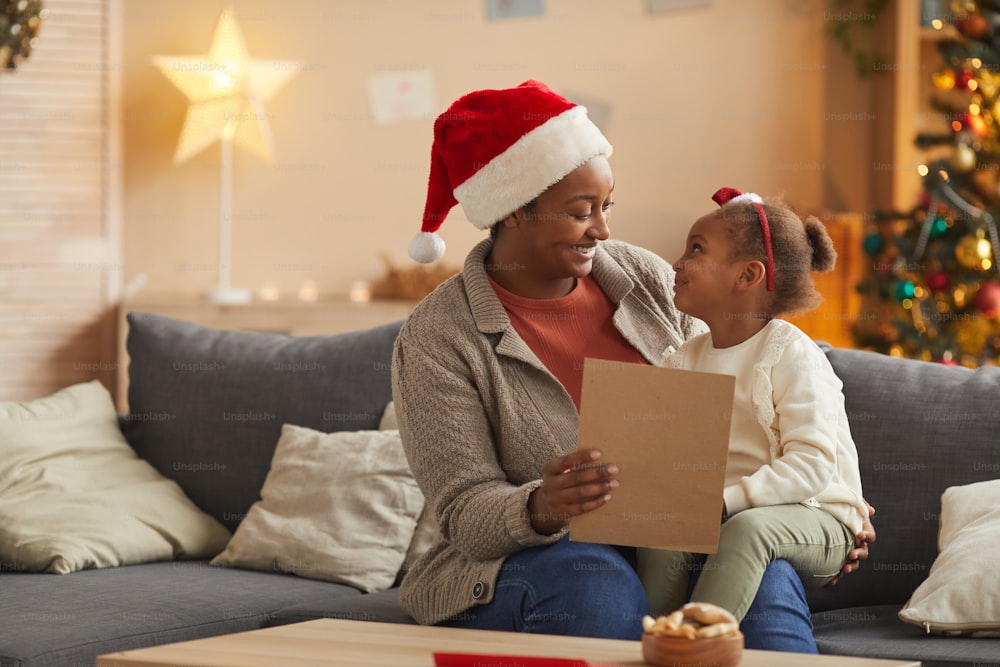 Ritratto della madre afroamericana sorridente che legge la lettera a Babbo Natale con la bambina carina mentre si gode il periodo natalizio a casa, spazio di copia