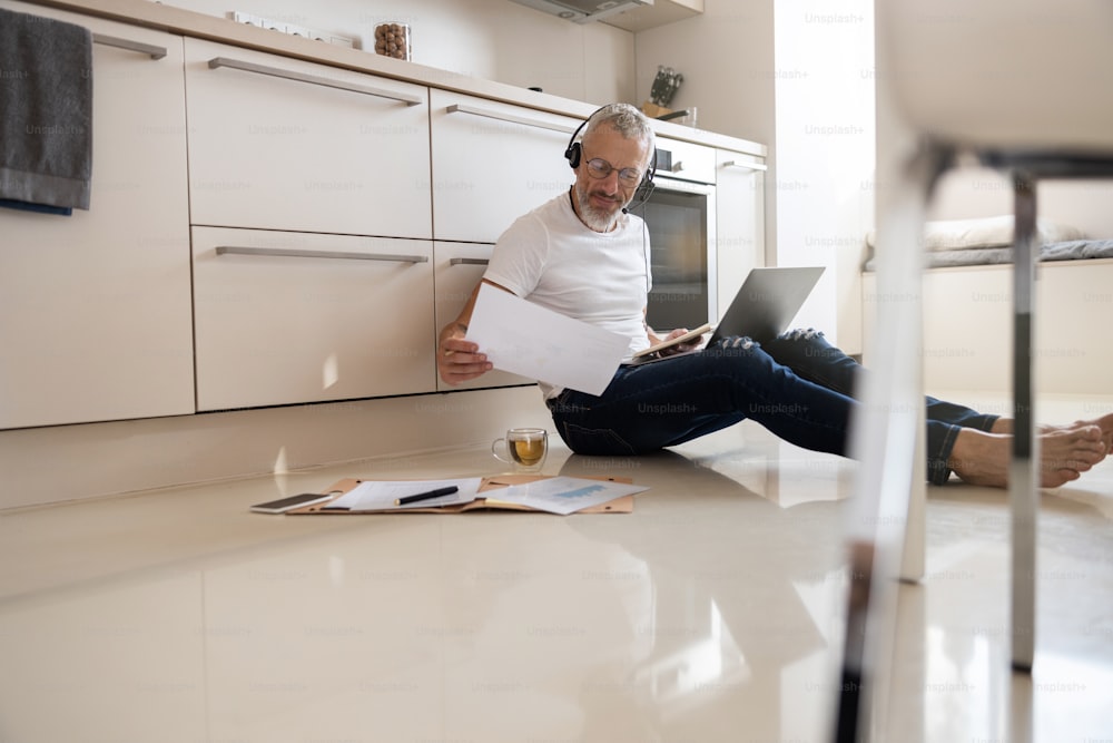 Spécialiste masculin travaillant à distance faisant ses recherches avec une pile de papiers et un ordinateur portable tout en s’appuyant sur une table de cuisine