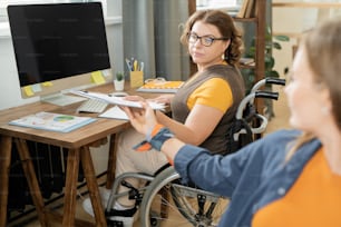 Junge ernsthafte Geschäftsfrau im Rollstuhl nimmt Klemmbrett mit Finanzdokument aus den Händen eines Kollegen während der Arbeit mit Daten im Büro