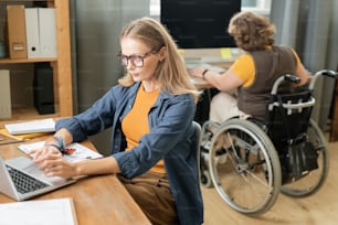 Giovane imprenditrice contemporanea che si concentra sul lavoro davanti al laptop durante l'analisi dei dati finanziari contro il collega disabile