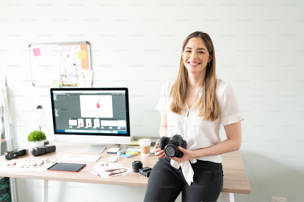 Femme photographe tenant un appareil photo et ayant l’air heureuse assise sur un bureau dans son studio