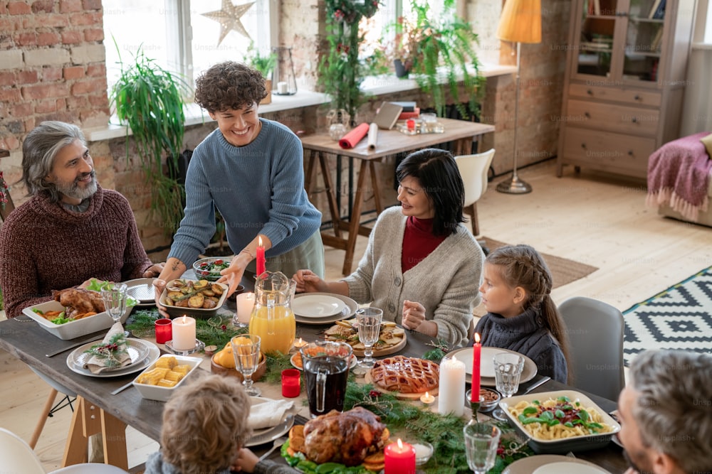 Joven y alegre mujer morena poniendo patatas asadas en la mesa festiva y mirando a su marido durante la cena familiar el día de Navidad