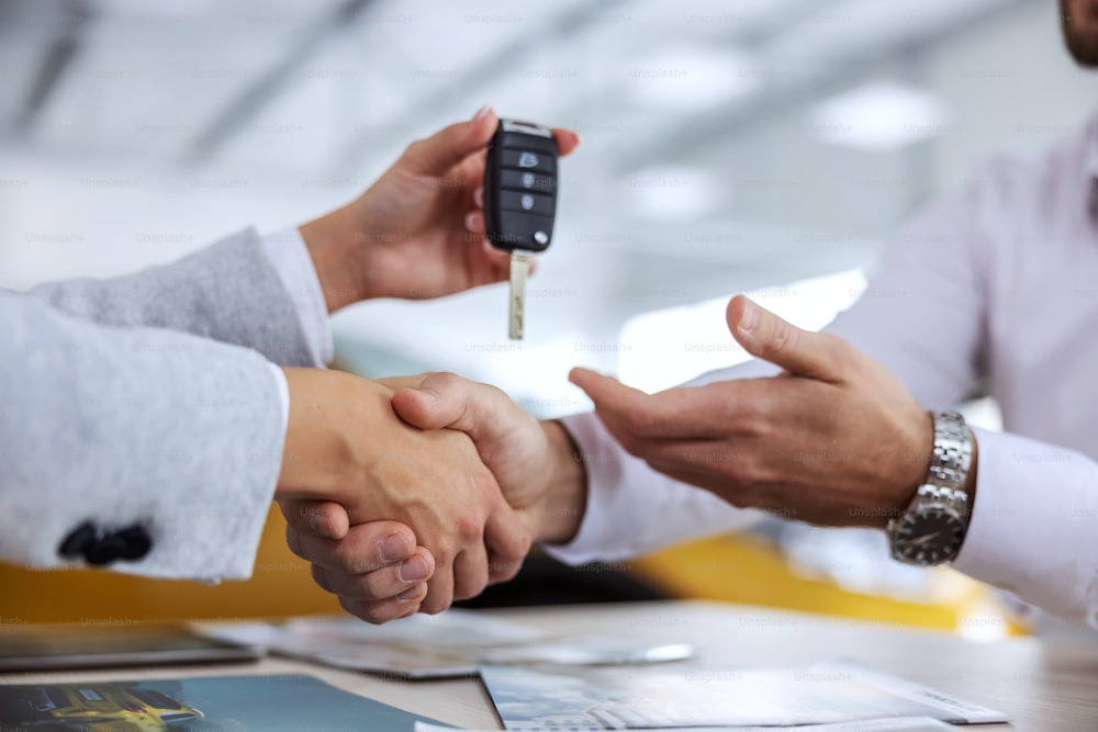 Primer plano del vendedor de automóviles y el comprador dándose la mano. Vendedor de automóviles entregando las llaves de un automóvil a un comprador. Interior del salón de coches.