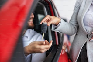Closeup of female car seller handing a car keys to a buyer who is sitting in new car. Car salon interior.