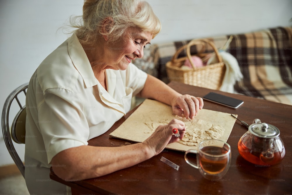 Smiling old lady creating male portrait on handmade picture while sitting at the table with glass teapot and cup of tea